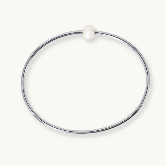 A minimalist polished silver-tone bangle bracelet with a single small white pearl bead at the top, shown on a clean off-white background.