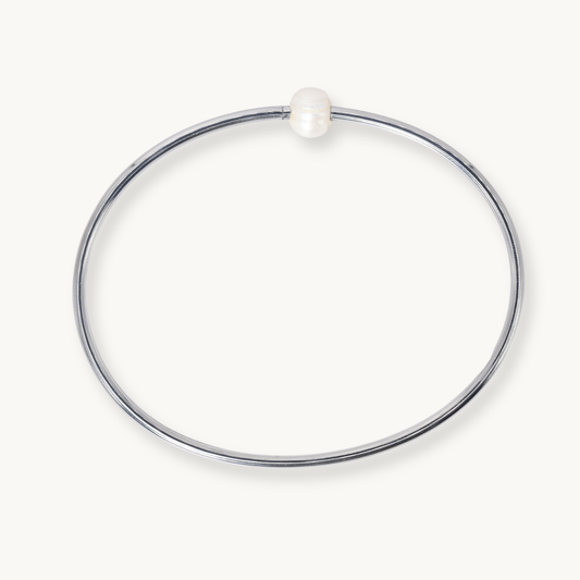 A minimalist polished silver-tone bangle bracelet with a single small white pearl bead at the top, shown on a clean off-white background.