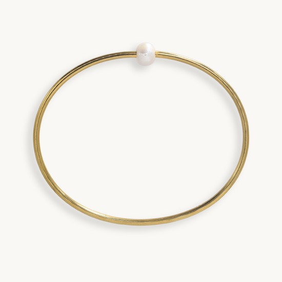A thin gold bangle bracelet with a single white pearl bead at the closure, shown on a clean off-white background.