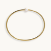 A thin gold bangle bracelet with a single white pearl bead at the closure, shown on a clean off-white background.