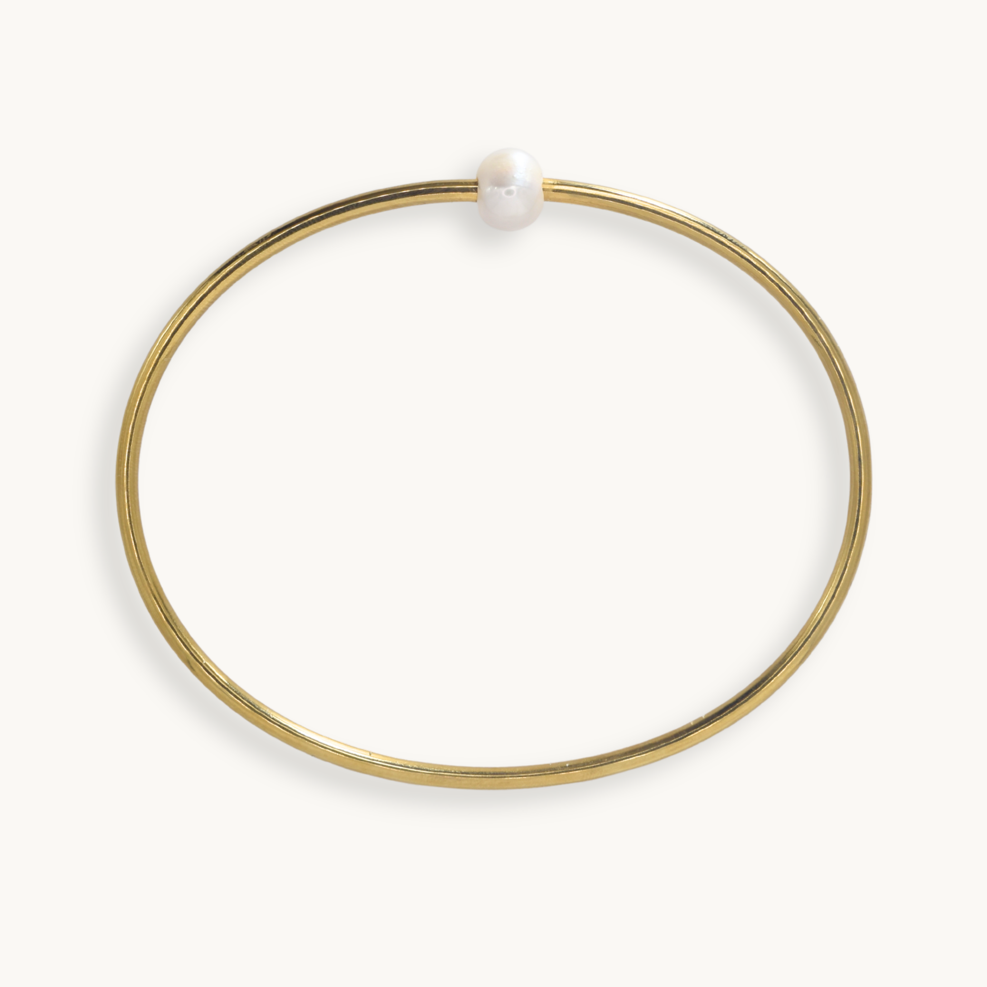 A thin gold bangle bracelet with a single white pearl bead at the closure, shown on a clean off-white background.