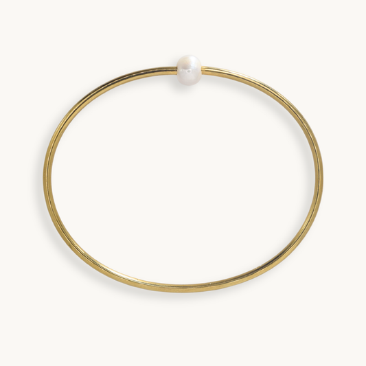 A thin gold bangle bracelet with a single white pearl bead at the closure, shown on a clean off-white background.
