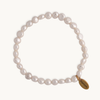 A delicate pearl bracelet with slightly irregular white pearls, strung on a clear thread. Features a small gold tag with engraved text, set against a plain light background.