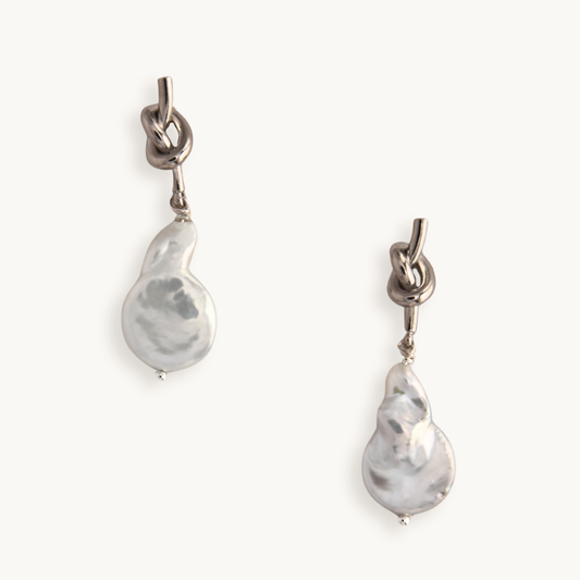 Silver earrings feature a unique knotted design with dangling baroque pearls, creating an elegant and artistic look against a plain white background.