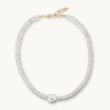 A delicate necklace made of small, tightly packed white beads with a single larger white shell pendant and a gold clasp, shown on a light cream background.