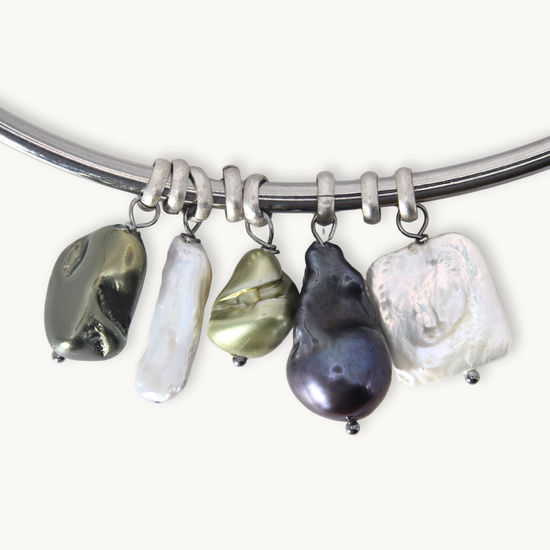 A silver-toned curved bracelet with five irregular baroque pearl charms in black, white, olive green, and dark purple, displayed on a light cream background.