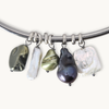 A silver-toned curved bracelet with five irregular baroque pearl charms in black, white, olive green, and dark purple, displayed on a light cream background.