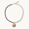 A beaded bracelet with a split design of dark brown and light pinkish-beige beads, featuring a gold-tone lobster clasp and a textured gold-tone sand dollar pendant, shown on a light cream background.