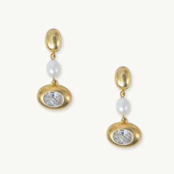 A pair of gold drop earrings featuring a polished oval gold top, a white pearl bead, and a larger oval gold setting holding a clear faceted gemstone, shown on a light cream background.