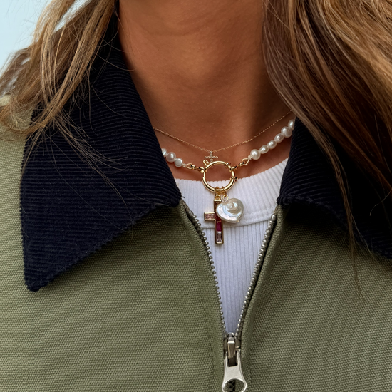 A close-up of a person wearing layered necklaces featuring a gold chain with a pearl heart and cross, and a pearl choker. They wear a green jacket.