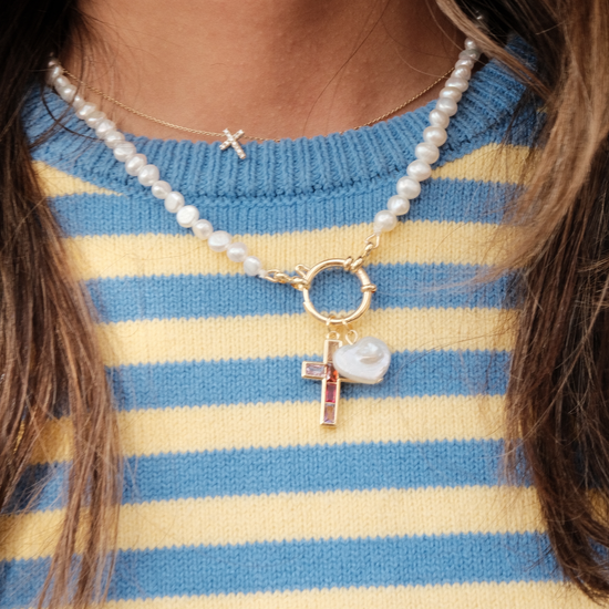 Close-up of a pearl necklace with a gold clasp, featuring a gemstone cross and a glossy heart charm. Worn over a blue and yellow striped sweater.