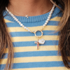Close-up of a pearl necklace with a gold clasp, featuring a gemstone cross and a glossy heart charm. Worn over a blue and yellow striped sweater.
