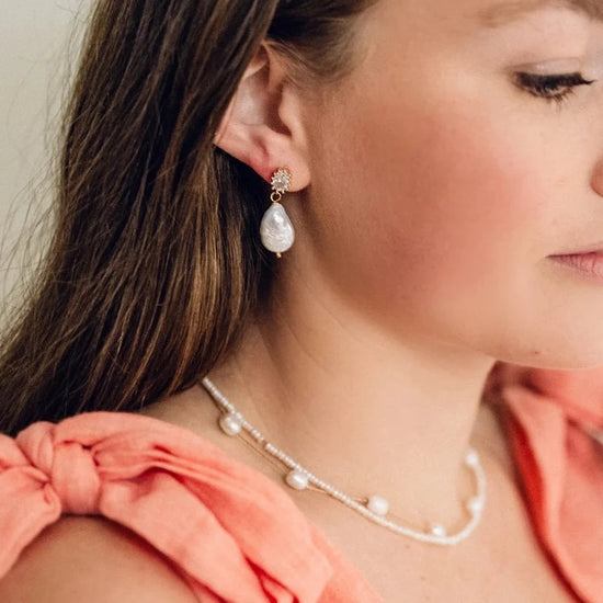 Woman wearing pearl drop earrings and a matching beaded necklace. Her peach top features a tied shoulder accent. The tone is elegant and serene.