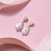 Elegant pearl drop earrings with gold accents and sparkling oval gems, set against a soft pink background, convey luxury and sophistication.