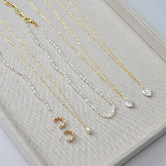 A beige display showcases elegant gold and pearl necklaces, with heart and drop pendants, and a pair of textured gold hoop earrings underneath.