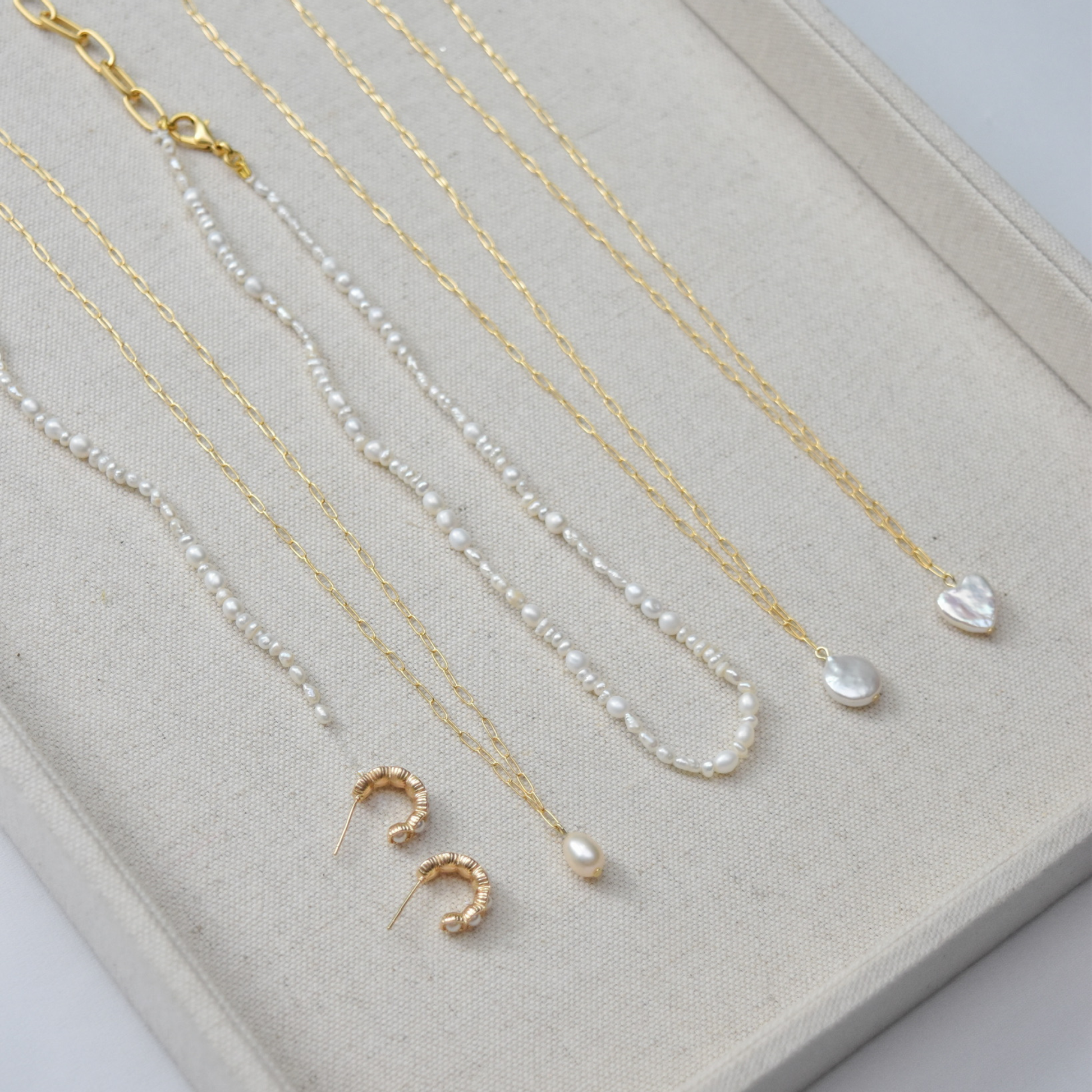A beige display showcases elegant gold and pearl necklaces, with heart and drop pendants, and a pair of textured gold hoop earrings underneath.