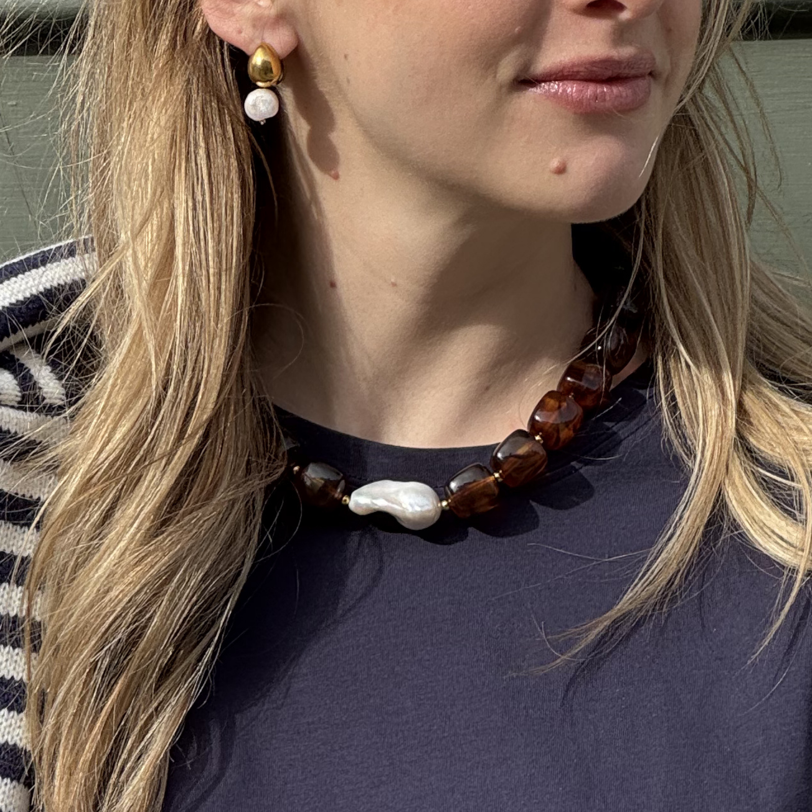 Woman wearing gold and pearl earrings, a chunky brown bead necklace with a large, irregular white pearl, against a navy top and striped sweater.