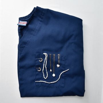 Folded navy blue scrub top adorned with gold and pearl necklaces draped over the front pocket, against a plain white background.