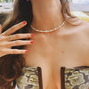 A woman with red nail polish and a gold ring touches a pearl necklace on her neck. She wears a strapless patterned top. The mood is elegant and relaxed.