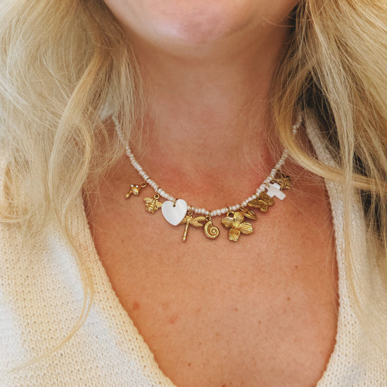 A person wearing a pearl necklace with gold charms, including a heart, flowers, and other shapes, set against a cream-colored knitted garment.