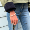 A hand wearing a pearl bracelet and silver ring rests against blue jeans, holding sunglasses. The casual outfit includes a green and navy jacket.