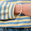 A person's wrist wearing a delicate pearl bracelet with a gold clasp, set against a cozy blue and yellow striped sweater, conveying a casual elegance.