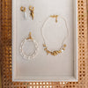 Elegant jewelry set on beige fabric includes gold hoop earrings with charms, a pearl bracelet, and a pearl necklace with gold charms.