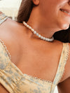 A woman wearing a pearl necklace and a floral yellow dress, smiling slightly. The mood is warm and relaxed, with soft lighting enhancing the warm tones.