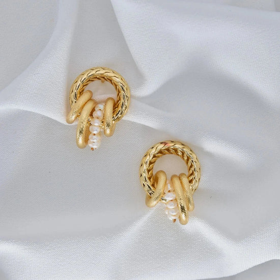 Gold hoop earrings with textured, braided design and small white pearls placed on soft, white fabric, conveying elegance and luxury.