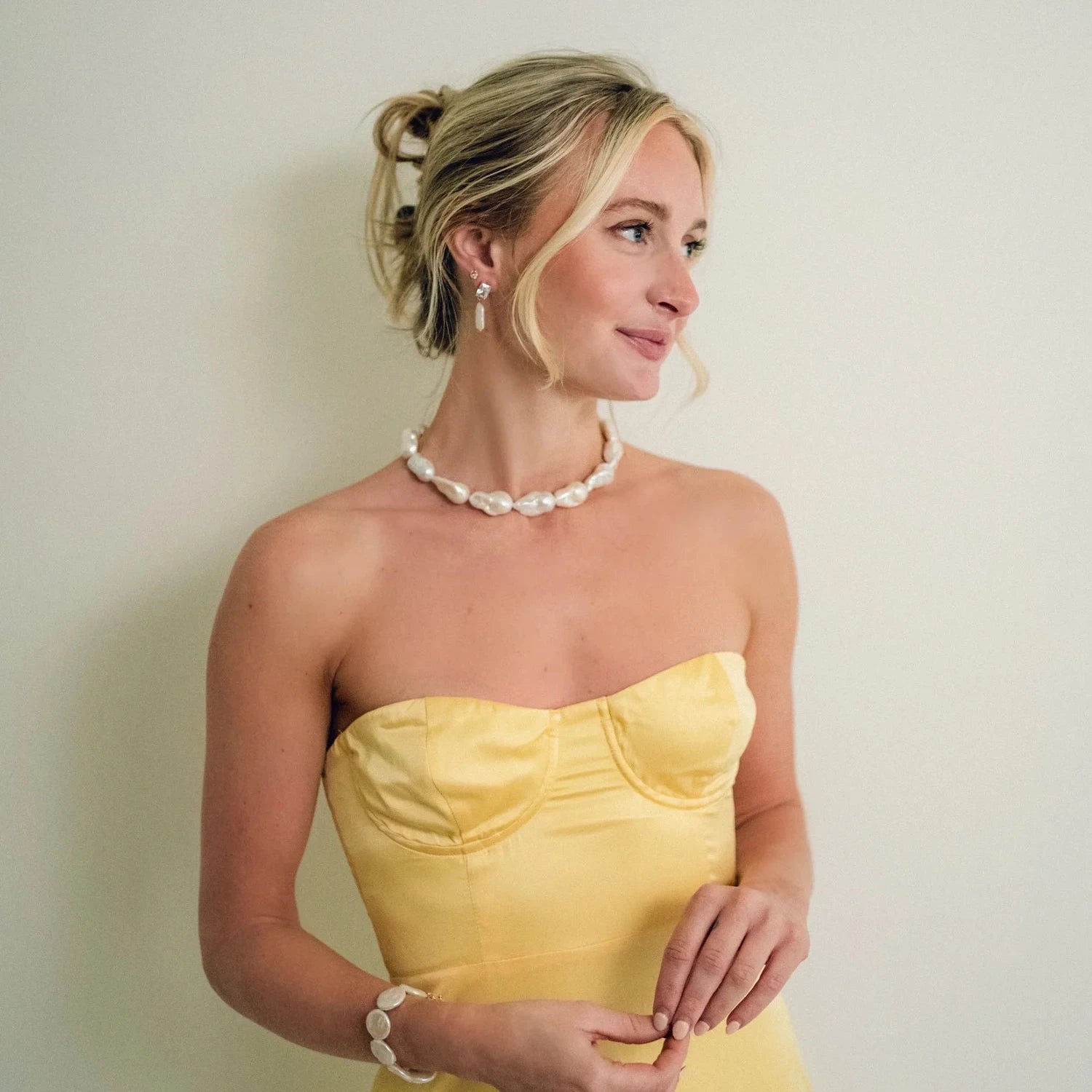 A woman in a strapless yellow dress smiles gently, wearing a pearl necklace and earrings. Her hair is styled in a loose updo, exuding elegance and grace.